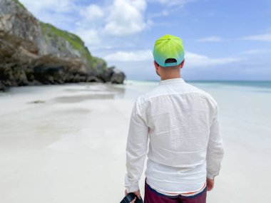 Zanzibar 'daki okyanusun beyaz kumlu sahilinde yürüyen şapkalı genç bir adam. Egzotik bir ülkeye gitmek.