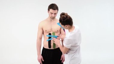 Kadın terapist beyaz arka planda erkek karnına kineziyoloji bandı uyguluyor. Kadın erkek hastayı Kinesio yapışkan bantla yapıştırmaya hazırlıyor.