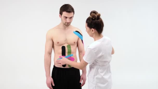 Female therapist applying kinesiology tape on a mans abdomen on the white background. Woman prepares male patient to glue kinesio adhesive tape — Stock Video