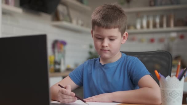 Умный мальчик использует ноутбук для видеосвязи со своим учителем. Экран снимает онлайн-лекцию с учителем, вытаскивающим предмет из комнаты, мальчик пишет вниз информацию. — стоковое видео