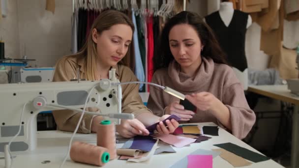 Två kvinnliga modedesigners tittar på prover av skisser i en syverkstad studio. Två skräddare eller sömmerskor som arbetar med symaskin och tygprover — Stockvideo