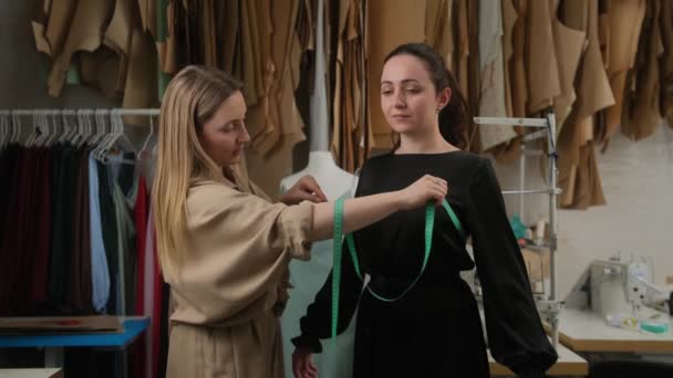 Kadın terzi atölyede bant kullanarak kadın müşterinin ölçümlerini alıyor. Terzilik stüdyosunda birlikte çalışan iki kadın. Tasarımcı ve model elbise ölçüleri için moda gösterisine hazırlanıyor. — Stok video