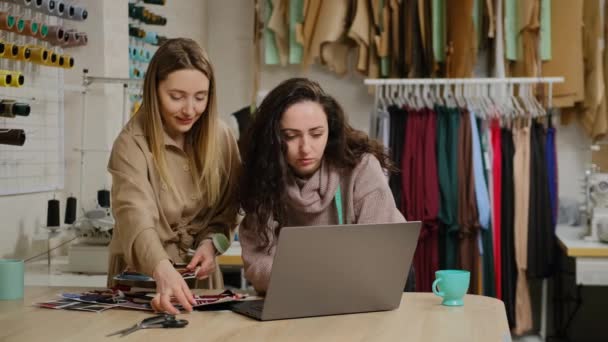 Colleghi sarto femminile guardando lo schermo del computer portatile, toccando campioni di tessuto e discutendo i colori mentre lavorano insieme in studio di cucito — Video Stock
