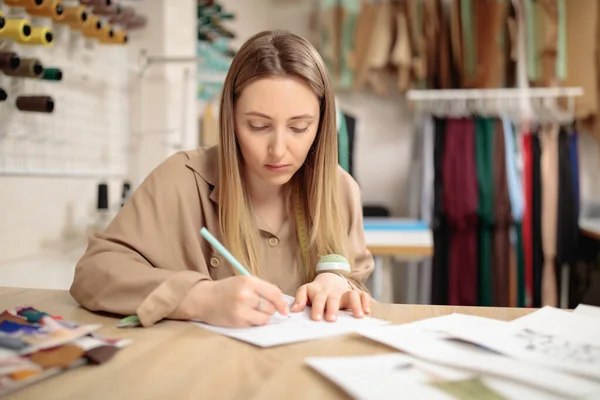 Young female fashion designer drawing sketch with model sitting in her office atelier.