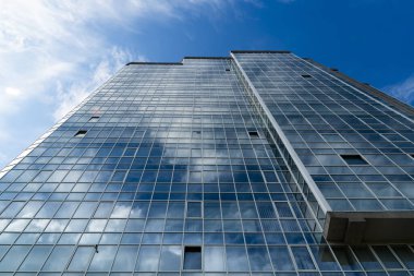 Uzun, modern bir cam ve çelik gökdelenin alçak açılı görüntüsü. Mavi gökyüzü ve beyaz bulutlar birçok penceresine yansıyor.