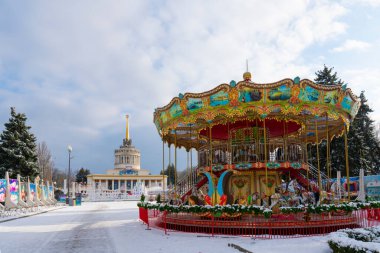 Kyiv, Ukrayna: 02.15.2025: Karlı bir kış parkı ortamında, tarihi bir sergi pavyonu ve arka planında altın kulesi bulunan canlı, süslü bir Noel atlıkarınca