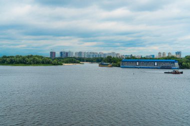 Önünde mavi yüzen bir otel ya da mavna bulunan geniş bir nehir manzarası ve uzak kıyılarda modern bir yerleşim yeri ve inşaat vinçleri.