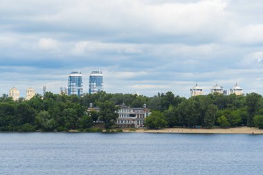 Geniş bir nehir manzarası kumlu bir sahil, yemyeşil ağaçlar ve dramatik bulutlu bir gökyüzünün altında modern gökdelenler.