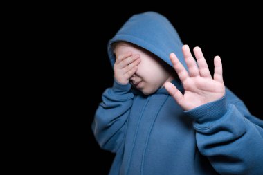 Mavi kapüşonlu bir kazak giyen küçük bir çocuk kameraya el hareketi yaparken utançla ya da korkuyla gözlerini kapatıyor.