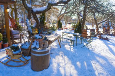 Karlı bir kış bahçesinde, rahat bir kafeterya terasında ahşap masalar, sandalyeler, beyaz buz patenleri ve parlak güneş ışığı altında sallanan bir at.
