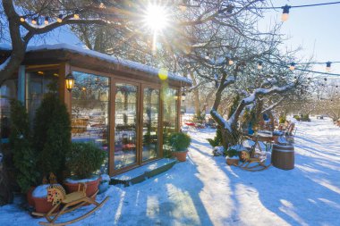 Parlak güneş parlaması, sallanan atlar ve açık hava tel ışıklarıyla kaplı, karla kaplı, rahat bir bahçe kafesi. Güneşli bir parkta güzel bir kış terası