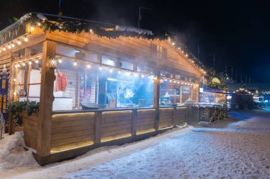 Açık hava kış tatili pazarında parlayan ışıklarla süslenmiş rahat ahşap bir büfe. Karlı bir akşamda sıcak yiyeceklerden buhar yükselir.