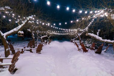 Tahta adirondack sandalyeler karlı bir patika boyunca sıcak ip ışıklarıyla dekore edilmiş bir bahçede. Bahçede kış tatillerinin büyülü akşam atmosferi