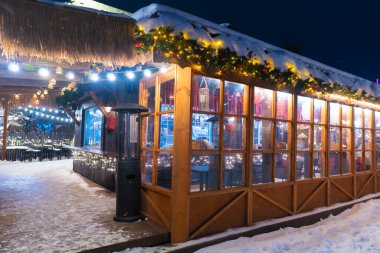 Açık hava kış tatili pazarında parlayan ışıklarla süslenmiş rahat ahşap bir büfe. Karlı bir akşamda sıcak yiyeceklerden buhar yükselir.