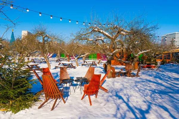 Karla kaplı bir kış bahçesinde, berrak mavi gökyüzünün altında, renkli sicim ışıkları olan modern ahşap şezlonglar. Açık havada güneşli bir gün.