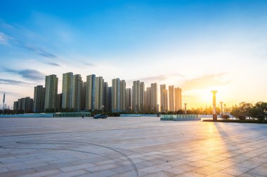 Boş asfalt yol, ağaçlar ve güneş ışığı altında gökdelenler.