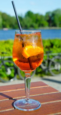 İki bardak parlak turuncu Aperol Spritz. Caddedeki bir kafede, bir nehrin ve yeşilliğin arka planında..