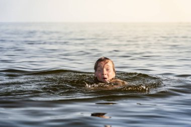 Neşeli bir çocuk sıcak bir yaz akşamı sakin bir gölde yüzer. Çocuk su sıçratırken geniş ölçüde gülümser, doğrudan kameraya heyecanla bakar..