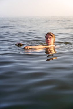 Genç bir çocuk, sakin göl suyunda gözleri kapalı bir şekilde uzanıyor ve huzurlu bir yaz akşamı güneşinin sıcağının tadını çıkarıyor..