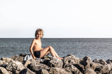 Genç bir çocuk su kenarındaki kayalara yerleştirilmiş bir havluya oturur. Güneşli bir yaz gününde, mayo giyerken ve manzaranın tadını çıkarırken kameraya gülümsüyor..