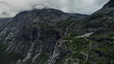 280 metre yüksekliğindeki Trollfossen şelalesi ve Storgrovfjellet dağını gösteren dik kayalıkların yanından inen insansız hava aracı. Norveç 'in doğal ortamındaki engebeli araziye giden yol boyunca bulutlar tepeleri kaplıyor.