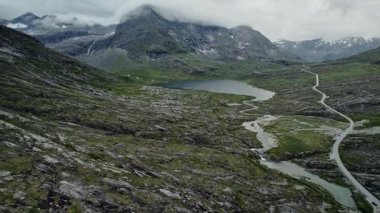 Sinema insansız hava aracı Norveç 'in Trollstigen kenti Alnesvatnet Gölü yakınlarındaki engebeli İskandinav arazisi boyunca yalnız bir virajlı yolu geri çekiyor. Görkemli dağlar, yeşil tundra ve vahşi doğa.