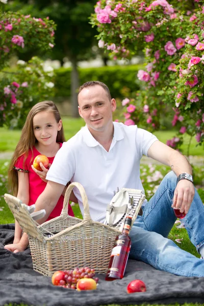 Hermoso padre joven sentado en un cuadros en un parque verde con su pequeña hija bonita con una canasta de mimbre para un picnic feliz — Foto de Stock