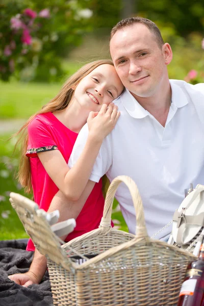 Hermoso padre joven sentado en un cuadros en un parque verde con su pequeña hija bonita con una canasta de mimbre para un picnic feliz — Foto de Stock