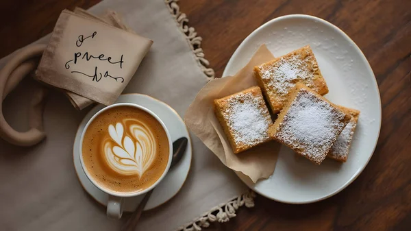 AI coffee and dessert concept showing a cozy caf setup with a cup of latte art coffee and a plate of square cakes dusted with powdered sugar.