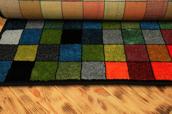 A multi-colored soft, rolled-up rug with a pattern of colored squares on a wooden floor. Fluffy carpet, interior element, flooring, floor covering