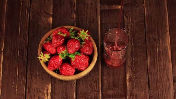 Red strawberry juice is poured into a glass and strawberry in a wooden bowl on a wooden table — Stock Video