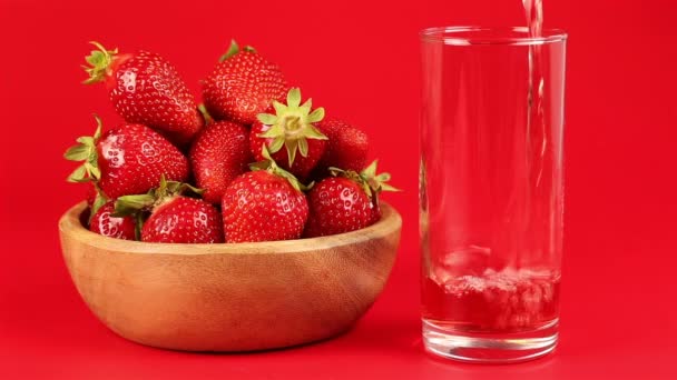 El jugo de fresa roja se vierte en un vaso y la fresa en un tazón de madera sobre fondo rojo — Vídeos de Stock
