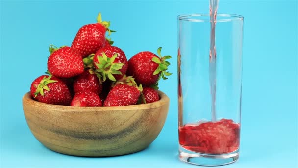 Красный клубничный сок наливают в стакан и клубнику в деревянной миске на голубом фоне — стоковое видео