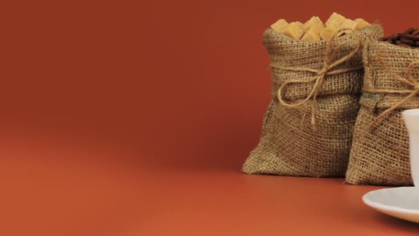 Cup of coffee. Behind stands coffee beans in burlap bag and next sugar in burlap bag on brown background — Stock Video