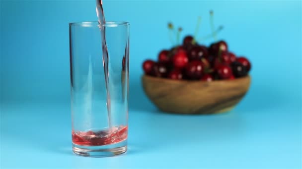 Suco de cereja vermelho sendo derramado em um copo e cerejas maduras vermelhas em tigela de madeira no fundo azul — Vídeo de Stock