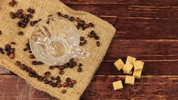 Coffee is poured into a glass cup on the table on him located coffee beans, burlap tablecloth and brown sugar cubes on brown wooden table — Stock Video