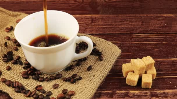 Kávé öntenek egy fehér csésze rá az asztalon található kávébab, a zsákvászon terítő és a barna cukor kockák barna fa tábla — Stock videók