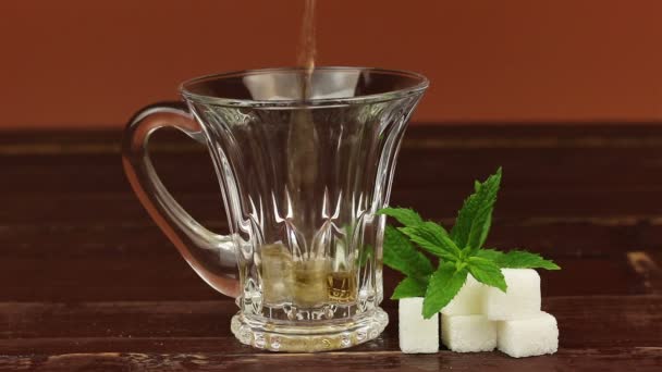 Thé versé dans une tasse à thé en verre et sucre blanc en cubes avec des feuilles de menthe sur une table en bois brun — Video