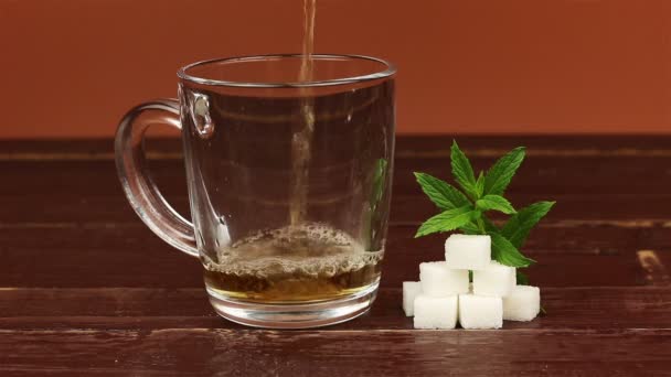 Té que se vierte en taza de té de vidrio y azúcar blanca en cubos con hojas de menta en mesa de madera marrón — Vídeos de Stock