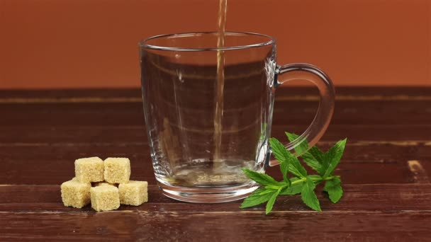 Tee in Glas Teetasse und braunen Zucker in Würfel mit Minzblättern auf braunem Holztisch gegossen — Stockvideo