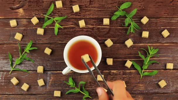 Кубики сахара добавляются в стакан для чая на столе со свежими листьями мяты и коричневым сахаром в кубиках на коричневом деревянном столе — стоковое видео