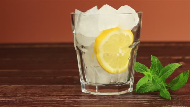 Vaso de té helado con menta y limón, cubitos de hielo sobre mesa de madera marrón — Vídeos de Stock