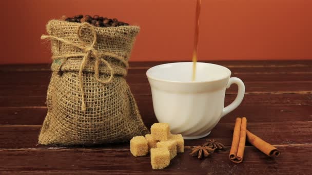 Despeje uma xícara de café na mesa localizada grãos de café em saco de serapilheira, cubos de açúcar mascavo e paus de canela sobre mesa de madeira marrom — Vídeo de Stock