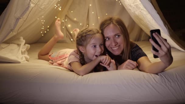 Gelukkig moeder en dochter worden gefotografeerd aan de telefoon in een geïmproviseerde tent in de woonkamer, ze glimlachen en lachen — Stockvideo