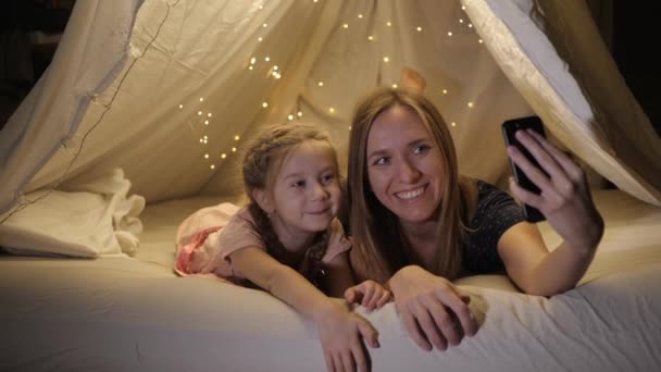Mama și fiica fericite sunt fotografiate la telefon într-un cort improvizat în camera de zi, zâmbesc — Videoclip de stoc
