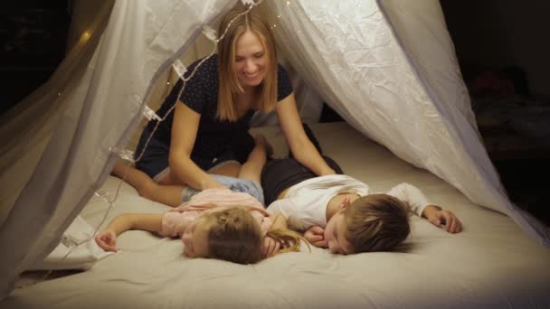 Glückliche Mutter genießt Familienurlaub mit ihren Kindern Sohn und Tochter im Zelt im Wohnzimmer, massiert die Kinder, legt sie ins Bett — Stockvideo