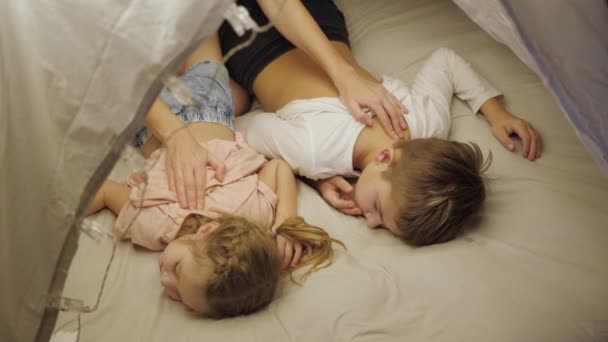 Feliz mamá disfruta de vacaciones familiares con sus hijos hijo e hija en una tienda de campaña en la sala de estar, masajes a los niños, los pone a la cama, la vista desde la parte superior — Vídeo de stock