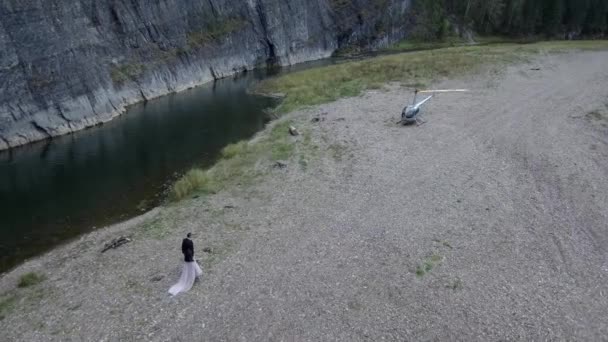 Una joven pareja toma de la mano y camina hacia su helicóptero, que está estacionado cerca del río bajo una roca. — Vídeo de stock