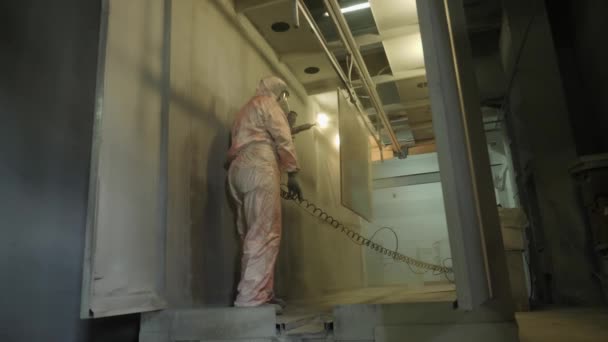 Работник в защитном костюме распыляет порошковую краску из пистолета на металлические изделия — стоковое видео
