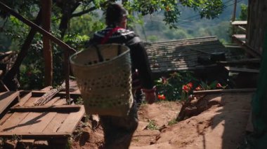 An Asian peasant woman and man carrying a wicker basket on their backs descend a dirt road in a traditional village setting - 4K Horizontal